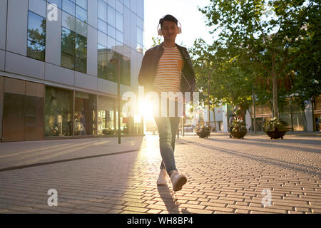 Portrait von lächelnden jungen Mann mit Kopfhörer zu Fuß auf der Straße an der Hintergrundbeleuchtung