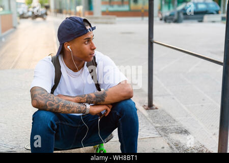 Tätowierte junge Mann sitzt auf seinem Skateboard Musik hören mit Smartphone und Kopfhörer.