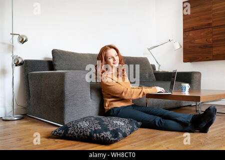 Die ganze Länge der Frau sitzt mit Laptop unterwegs im Wohnzimmer zu Hause Stockfoto