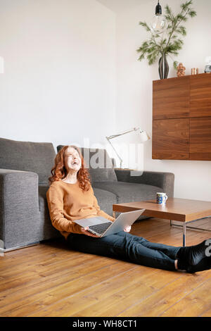 Die ganze Länge der fröhliche Frau lachend, während mit Laptop im Wohnzimmer zu Hause sitzen Stockfoto