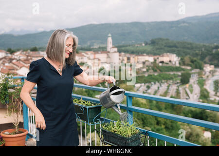 Lächelnde ältere Frau, Bewässerung von Pflanzen auf ihrer Dachterrasse, Belluno, Italien Stockfoto