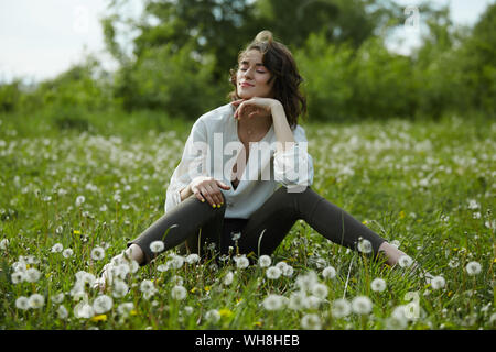 Frühling Portrait eines Mädchen sitzen in einem Feld auf dem Gras unter den Löwenzahn Blumen. Fröhliches Mädchen genießt sonnige Wetter im Frühling. Natürliche Schönheit einer woma Stockfoto