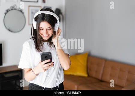 Glückliche junge Frau mit Smartphone und Kopfhörer zu Hause Stockfoto