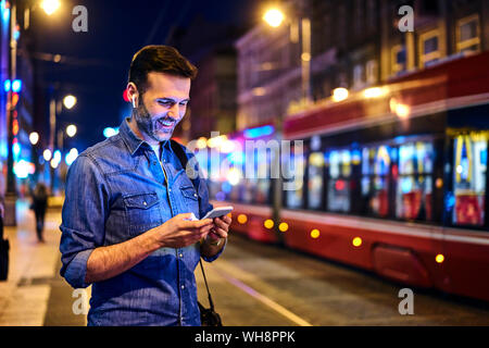 Lächelnder Mann mit drahtlosen Kopfhörern über Smartphone, während für die Straßenbahn bei Nacht warten Stockfoto
