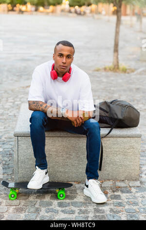 Portrait von tätowierten jungen Skateboarder sitzt auf der Bank mit Smartphone, Kopfhörer und Rucksack Stockfoto