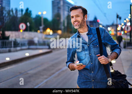 Lächelnder Mann mit drahtlosen Kopfhörern und Smartphone warten an der Straßenbahnhaltestelle am Abend nach der Arbeit pendeln Stockfoto