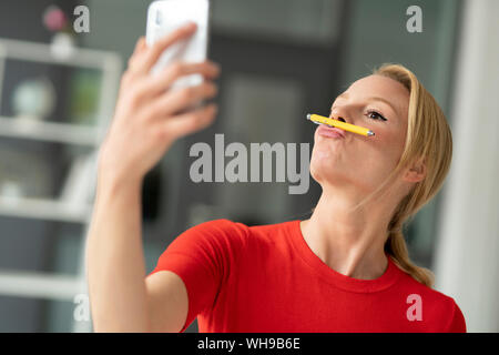 Verspielte junge Frau balancing Pen auf den Mund, im Büro ein selfie Stockfoto