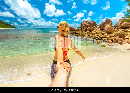 Blond touristische Frau im Bikini die Hand des Partners im Anse Caiman Strand mit natürlichen Pool im La Digue, Seychellen, Indischer Ozean, Afrika Stockfoto