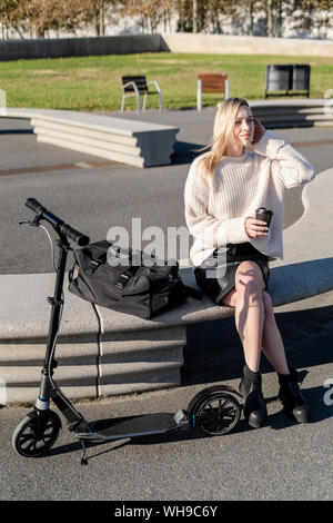 Blonde junge Frau mit Kick scooter sitzt auf der Bank mit Sporttasche Kaffee trinken zu gehen Stockfoto