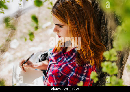 Nachdenklich, rothaarige Frau lehnte sich gegen Baumstamm Notizen Stockfoto