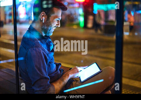 Mann mit Kopfhörer an einer Station sitzen in der Nacht mit seinem digitalen Tablet Stockfoto