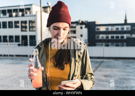 Stilvolle Junge Frau mit Getränk, Ohrhörer und Handy auf dem Parkdeck Stockfoto
