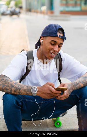 Portrait von tätowierten jungen Mann sitzt auf Skateboard Musik hören mit Smartphone und Kopfhörer. Stockfoto