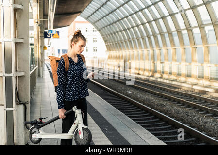 Junge Frau mit Elektroroller und Handy am Bahnhof Stockfoto