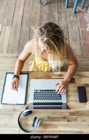 Junge Frau an den hölzernen Schreibtisch mit Laptop und Schreiben über die Zwischenablage Stockfoto