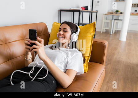 Junge Frau auf der Couch liegen zu Hause mit Smartphone und Kopfhörer Stockfoto