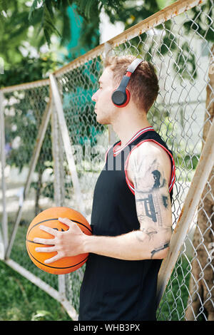 Schönen sportlichen jungen Mann lehnte sich auf netto rund um street Basketball Court und das Hören von Musik über Kopfhörer bei Bruch zwischen Viertel Stockfoto