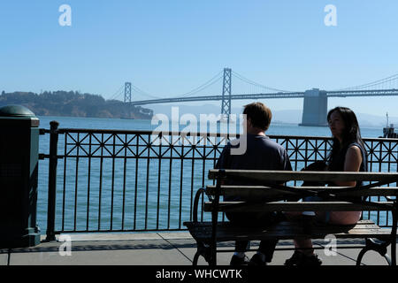 Junges Paar auf einer Bank sitzen mit Blick auf die Bay Bridge in San Francisco, Kalifornien an einem sonnigen Tag. Stockfoto
