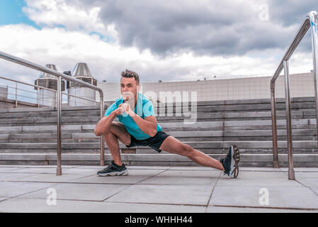 Athlet Mann tut das Bein strecken, bevor Morgen Workout. Im Sommer in der Stadt, Vorbereitung für einen Lauf, sportswear Shorts T-Shirt und Sneakers. Ste Stockfoto