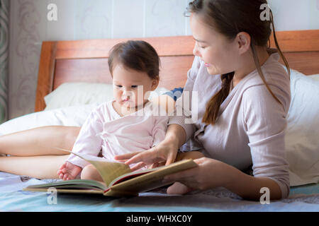 Eine Mutter eine Geschichte liest, die ihr Baby auf ein weiches Bett. Kind auf ein Buch konzentrieren. übergeordneten spiegeln die Seiten eines Bücher. homeschooling. Stockfoto