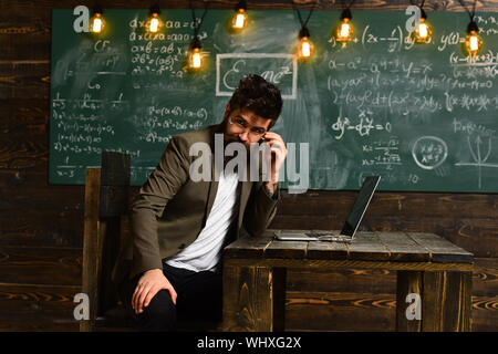 Bärtige Geschäftsmann mit Brille und Laptop sitzen und tippen, schwarzes Brett, die Glühbirnen im Hintergrund. Stockfoto