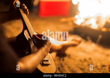 A happy Camper Spielen einer hawaiianischen Instrument Gitarre rund um den Campingplatz Feuer am Strand bei Nacht. Stockfoto