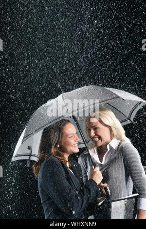 Zwei glückliche Geschäftsfrauen stehen unter einem Dach zusammen im Regen Stockfoto