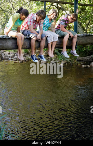 Happy Teenager Jungs und Mädchen sitzen auf Holzbrücke bei Stream nach unten schauen. Stockfoto