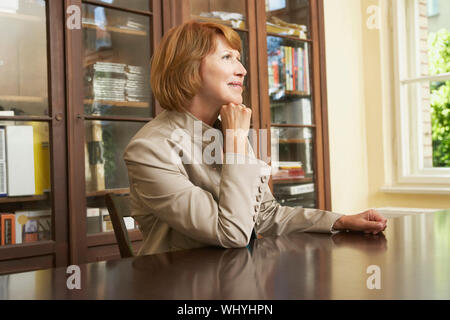 Seitliche Sicht auf eine Frau mittleren Alters an Studie Tisch sitzen im Wohnzimmer Stockfoto