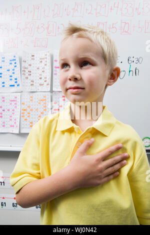 Little Boy in Gelb casual t-shirt weg schauen mit Hand auf Herz steht im Klassenzimmer Stockfoto