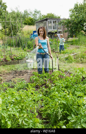 Glückliche Frau Bewässerung von Pflanzen in Grundstück mit Sohn im Hintergrund