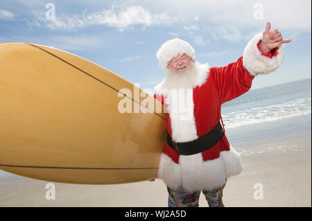 Happy senior Santa Claus und gestikulierte mit Surfbrett am Strand. Stockfoto