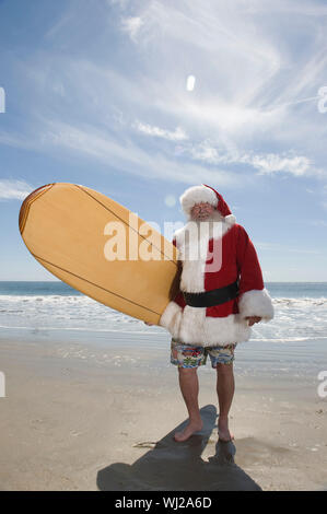 Volle Länge des Santa Claus mit Surfbrett am Strand. Stockfoto