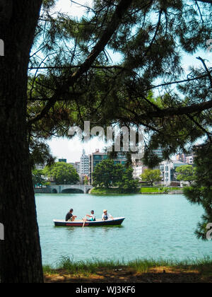 Menschen reiten kleines Boot in großen Körper von Wasser in Fukuoka, Japan von Natur Bäume mit städtischen Gebäude im Hintergrund umgeben Stockfoto