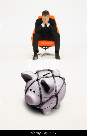 Besorgt Geschäftsmann auf einem Stuhl und Sparschwein mit Seil, die finanziellen Schwierigkeiten gebunden Stockfoto