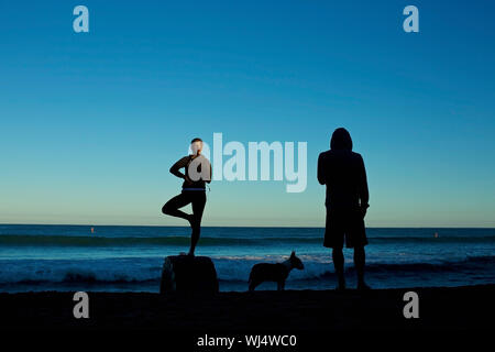 Silhouette junges Paar mit Hund Üben Yoga auf Sunrise Beach Stockfoto