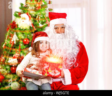 Santa Claus mit Enkelin sitzt neben Weihnachtsbaum, niedliche Mädchen öffnen Geschenk-Box, leuchtende Lichter, magische Nacht, Märchen-Konzept Stockfoto