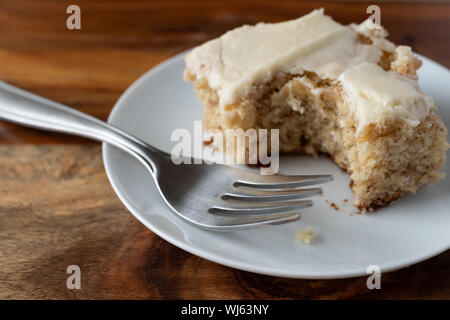 Seitliche Sicht auf die selbstgemachte Banane Brot mit Vanille Glasur gebissen auf einem Teller mit der Gabel auf einem Holztisch mit natürlichem Licht beleuchtet. Stockfoto