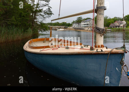 Ein kleines Segelboot mit einem blauen Rumpf schwimmend auf einer Wasserstraße mit Häusern im Hintergrund. Stockfoto