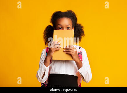 Schwarze Schülerin versteckt sich hinter Buch über gelben Hintergrund Stockfoto