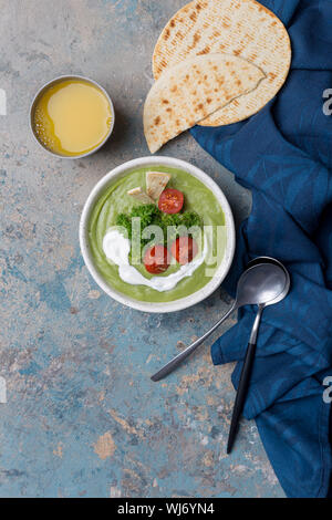 Grüne Cremesuppe mit Sahne und Brokkoli und Tomaten auf Beton. Blau serviette, Löffel, Pita und Orangensaft in der Nähe. Konzept der saisonale Gerichte. Umhüllung Stockfoto