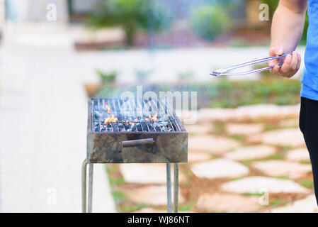 Schöner Athlet männlichen bereitet Grill im Freien. Brutale bärtiger Mann stehen und bereit, Grill, Holzkohle outdoor mit Sonnenuntergang Natur Sommer Garten Hintergrund brennen. Stockfoto