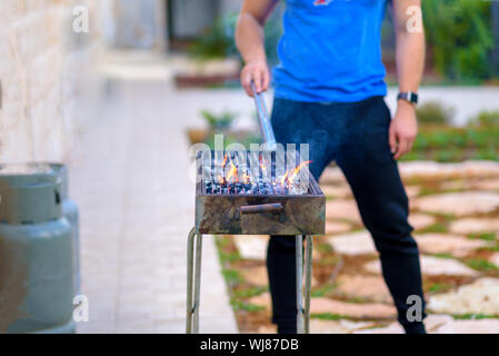 Schöner Athlet männlichen bereitet Grill im Freien. Brutale bärtiger Mann stehen und bereit, Grill, Holzkohle outdoor mit Sonnenuntergang Natur Sommer Garten Hintergrund brennen. Stockfoto