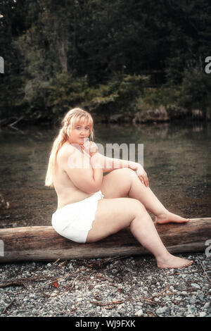 Junge curvy Frau sitzt leicht bekleidet auf einem See auf einem Baumstamm Stockfoto