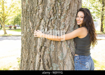 Casual wunderschöne Brünette mit einem Baum an der Kamera in einem Park an einem sonnigen Tag Stockfoto