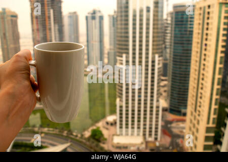 Weibliche Hand hält grosse weiße Tasse heißes Getränk und Wolkenkratzer der modernen Stadt Dubai mit kopieren. Kaffeetasse auf Stadt Hintergrund Stockfoto