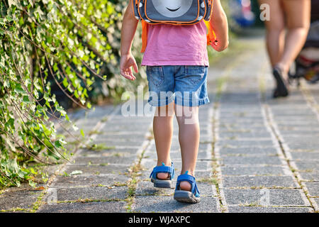 Ansicht von hinten von einem 3-4 jährigen Kind zu Fuß hinter seine Mutter mit einem Rucksack auf dem Bürgersteig an einem sonnigen Sommertag Stockfoto