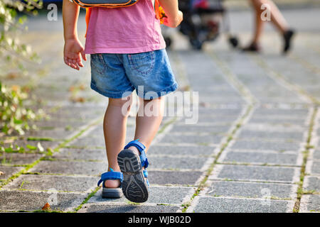 Ansicht von hinten von einem 3-4 jährigen Kind zu Fuß hinter seine Mutter mit einem Rucksack auf dem Bürgersteig an einem sonnigen Sommertag Stockfoto