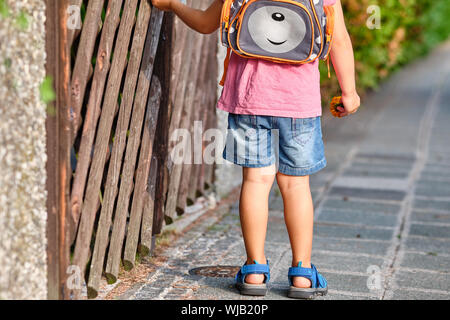 Ansicht von hinten von einem 3-4 jährigen Kind stehend an Holz rustikaler Zaun mit Rucksack auf dem Bürgersteig an einem sonnigen Sommertag Stockfoto