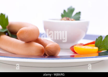Drei kalt geräucherte Würstchen oder Wiener Würstchen serviert auf einem Teller mit einem Sprung in eine kleine Schüssel geben und in Scheiben geschnittenen Kirschtomaten auf ein weißes Studio-Hintergrund Stockfoto